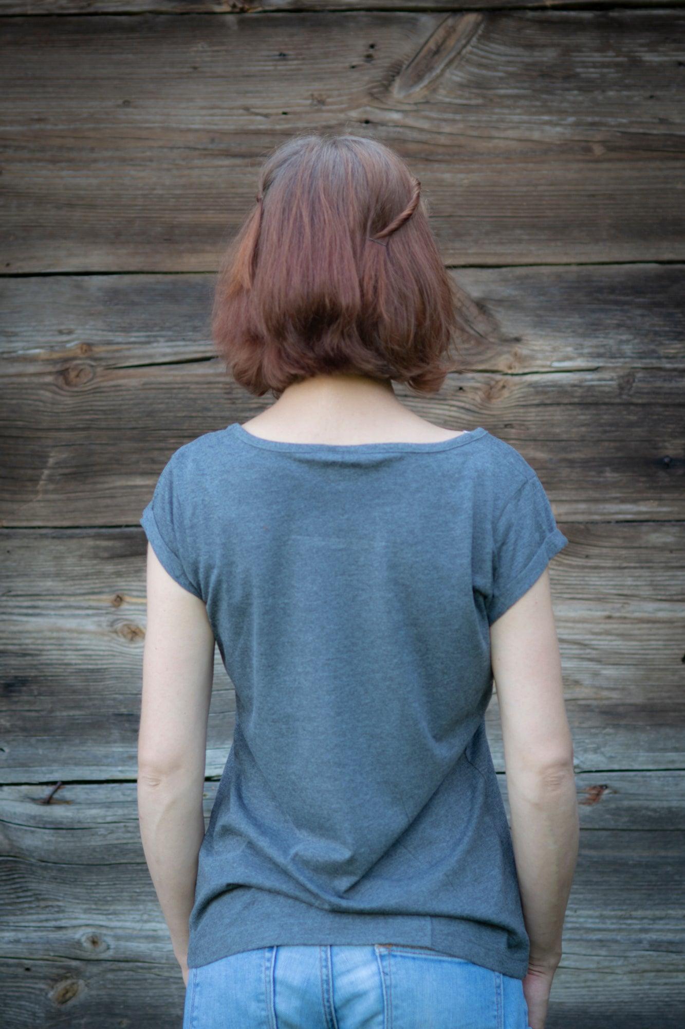 Meitene stāv pie koka mājas sienas pagriezusi muguru. Mugurā tai pelēks T-krekls uz kura sietspiedes tehnikā uzdrukātas piecas tautu meitenes baltā krāsā.