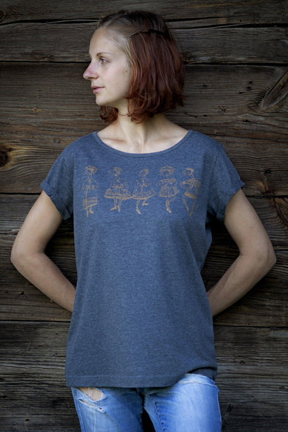 Meitene stāv pie koka mājas sienas. Mugurā tai pelēks T-krekls uz kura sietspiedes tehnikā uzdrukātas piecas tautu meitenes bronzas krāsā.