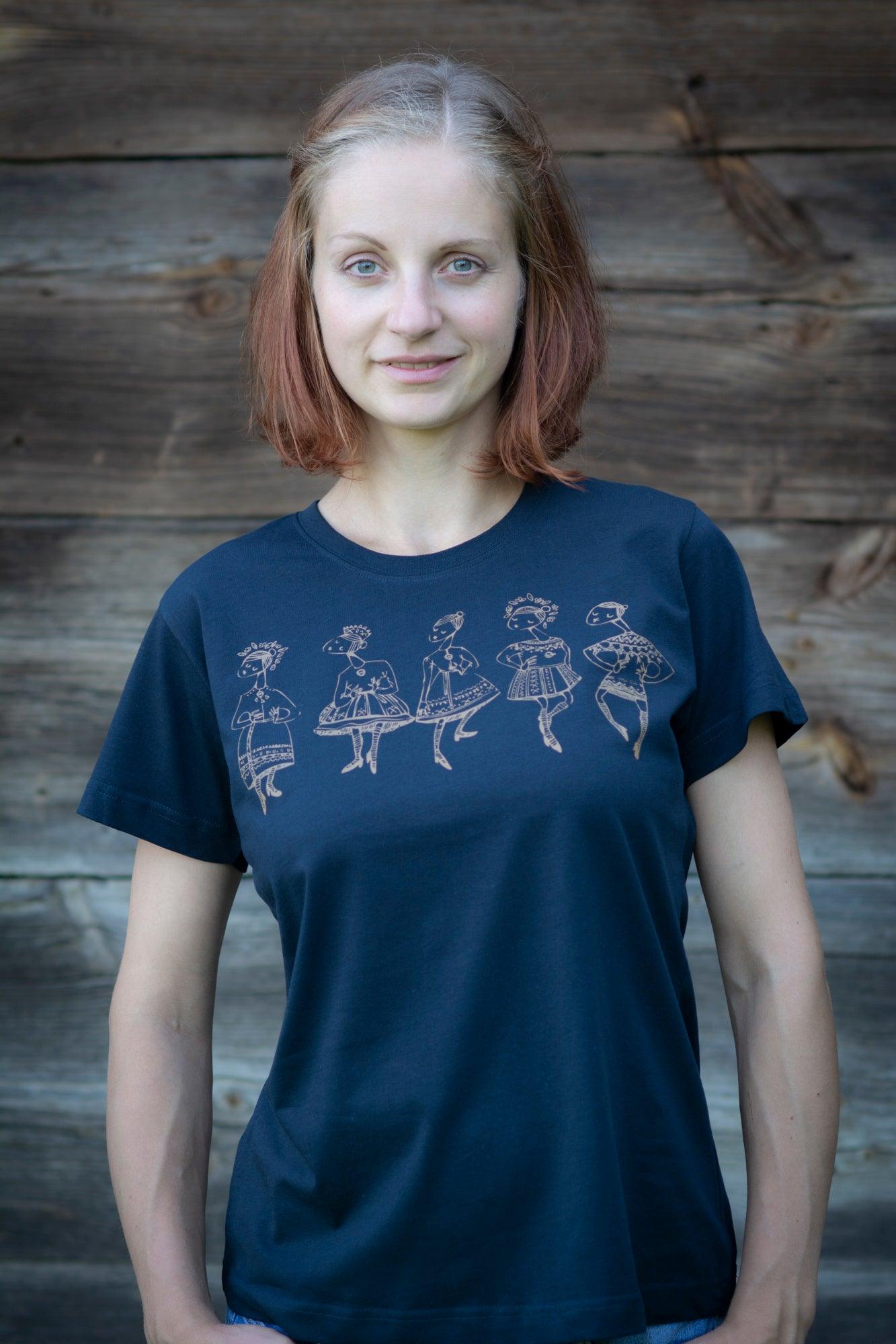Meitene stāv pie koka mājas sienas. Mugurā tai tumši zils T-krekls uz kura sietspiedes tehnikā uzdrukātas piecas tautu meitenes bronzaskrāsā
