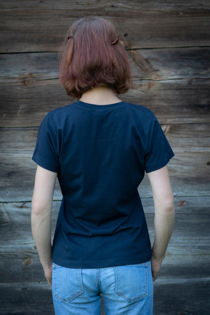 Meitene stāv pie koka mājas sienas pagriezusi muguru. Mugurā tai tumši zils T-krekls .